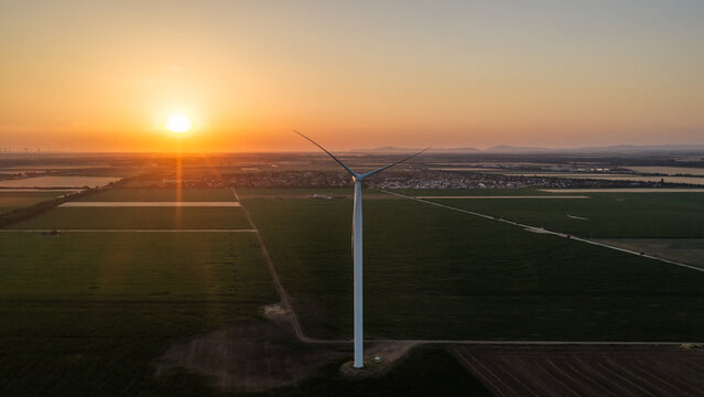 Aerial view of a wind turbine standing in a vast green agricultural field during a golden sunset with a clear sky in Level, Hungary.