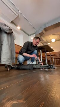 Low-angle view of a man kneeling on a wooden cafe floor as he inspects and tunes an electric guitar beside its open case. Warm pendant lights and curved