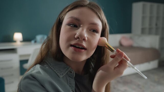 Teen girl applying makeup with brush in cozy bedroom, focused expression as she blends blush near cheekbone, denim shirt, warm lamp glow, soft teal wall, closeup beauty routine with natural skin