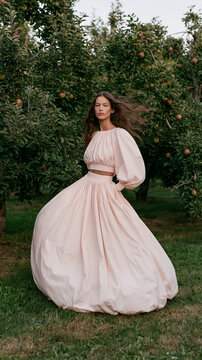 Elegante junge Frau in langem rosafarbenem Kleid steht in einem bl&uuml;henden Obstgarten. Romantisches Fashion-Portr&auml;t, feminine &Auml;sthetik, Natur, Fr&uuml;hling und vertr&auml;umter Lifestyle.