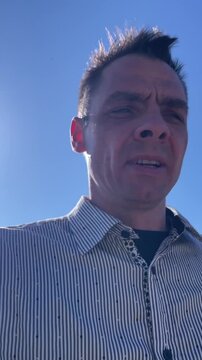 Tight low angle shot of a middle aged man framed on the right, cropped at chest level against a clear blue sky. Harsh midday sunlight creates strong shadows