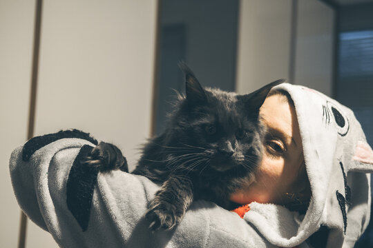 A young Caucasian woman with short hair holds a large black cat close to her face. She wears a cozy hoodie with animal patterns. The background is softly blurred.