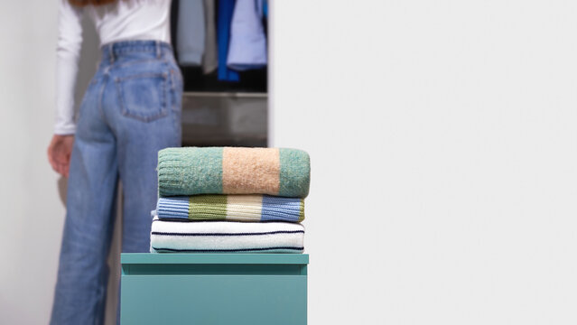 Folded colorful woolen sweaters with woman organizing open wardrobe on light gray background. Concept of closet organization, seasonal reset, clothing storage, sorting and decluttering. Copy space