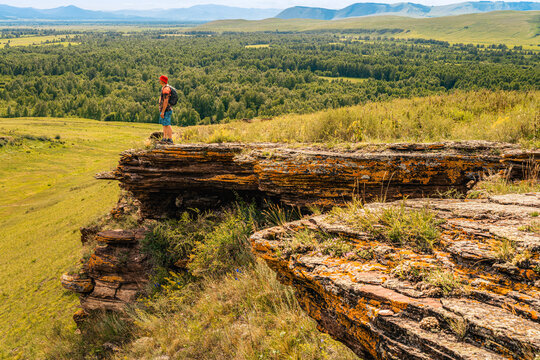 Hiker with backpack stands on edge of mossy cliff, admiring endless panorama of forest-steppe with mountains on horizon. A solo traveler without face, looking into distance, slow travel
