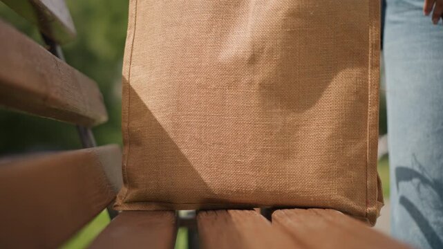 Tote bag resting on wooden bench, jeansclad person places jute tote on seat, closeup woven texture and bench slats, sunny urban park background, slow reveal emphasizing detail and composition