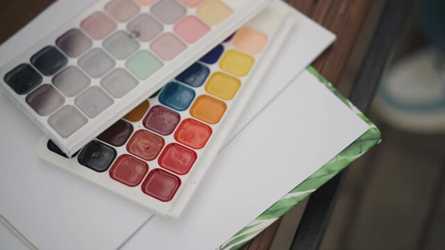 Watercolor palette and sketchbook resting on wooden tabletop, vibrant pigment pans, neat paper edge, soft natural light, closeup flatlay capturing artist workspace and creative tools