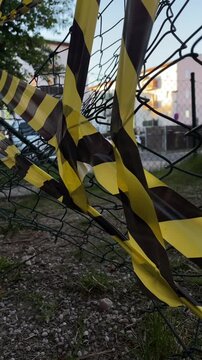 Yellow black tape fluttering across chainlink fence, maintenance crew surveys barrier near residential parking, cars and apartment facades in soft