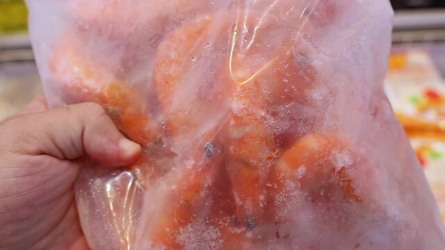 Frozen shrimp in plastic package held in customer hand at supermarket. Close up of icy orange prawns inside transparent bag, being selected by shopper from grocery store freezer display case area.