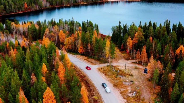 Red and gray cars navigating winding road surrounded by autumn trees and serene lake in Finland