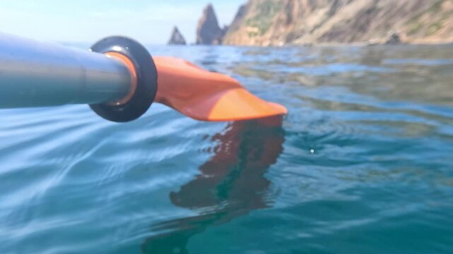 Kayak paddle ocean orange blade resting on calm turquoise water with a blurred scenic rocky coastline background for adventure travel