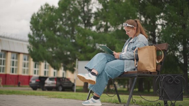White woman seated on bench writing notebook, denim outfit, sunglasses on head, tote bag beside, campus street and trees behind, concentrated expression, study vibe, handheld pencil closeups