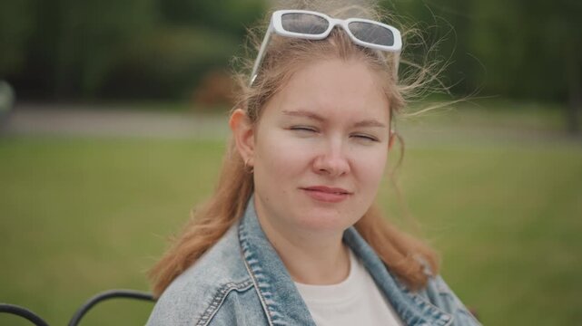 Caucasian woman sitting on park bench smiling softly wearing denim jacket and white sunglasses, late summer afternoon, soft bokeh background, natural light, gentle breeze ruffling hair, candid relaxed