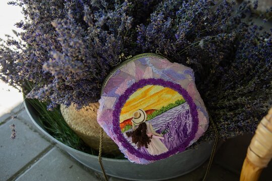 Handcrafted handbag adorned with vibrant embroidered landscape depicting a woman in lavender fields, set among fresh lavender flowers in a decorative metal container.