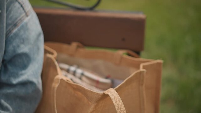 Closeup of tote bag and hand, hand of woman reaches into jute tote for water cup and brushes, denim sleeve and gingham cloth visible, bench and grass blurred in background, warm daylight texture study