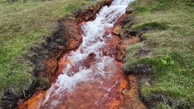 Small stream with iron-rich orange water flowing through grassy banks in a natural landscape, peaceful flowing water textures for environmental and geological themes
