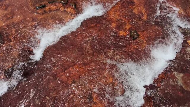 Top down overhead view of fresh clear water rushing over textured red rock riverbed with sparkling sun reflections and white foam