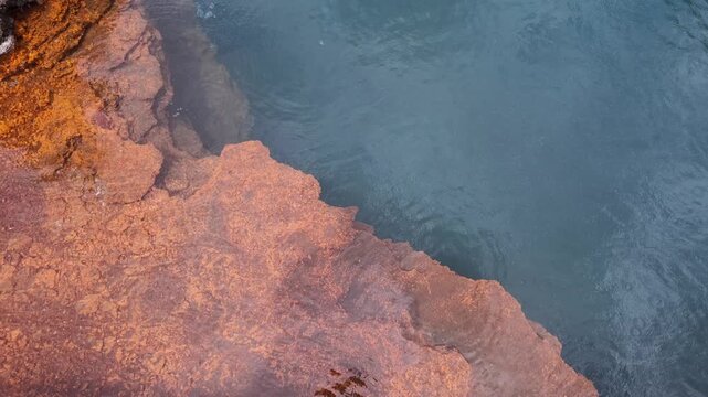 Top view of clear blue water flowing gently past textured orange rocky mineral deposits, natural thermal hot spring details, geological sulfur pool stream