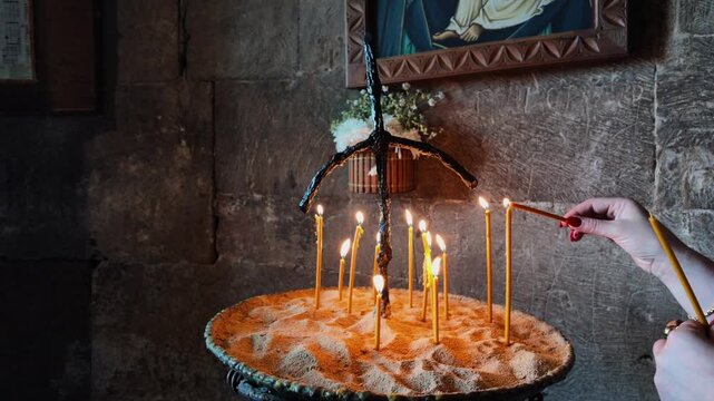 Woman lighting a thin wax candle from another flame in a dim stone church, faith and spiritual worship, christian religious tradition, solemn prayer atmosphere