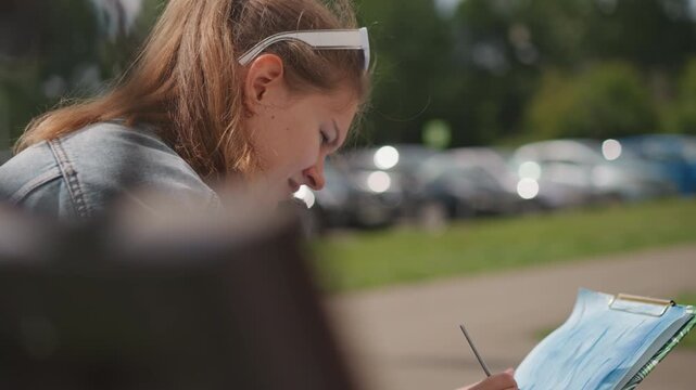 Young woman painting at bench near parking, denim jacket and ponytail, close profile view with brush and paper, cars in background bokeh, sunlight highlighting hair, focused hobbyist capturing color