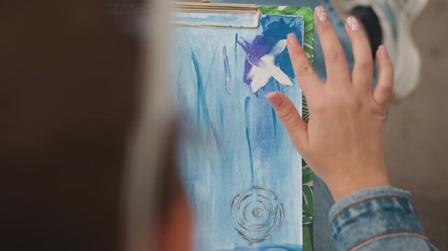 Artist finishing white detail on blue watercolor clipboard, finger forming starlike cross strokes, denim sleeve and sneakers visible, intimate closeup of tactile creation and final flourish, joyful