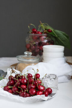 Close-up of a stack of Fresh cherries on parchment, cherries in a glass jar and a jar of brown sugar on a table