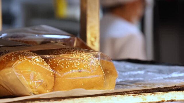 Sesame seed buns in plastic packaging displayed on a bakery shelf. Close up view of golden-brown baked bread rolls with sesame seeds ready for sale or sandwich making in a food shop.