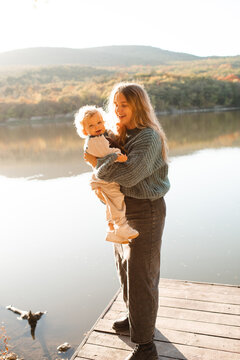 Young mother with little baby on wooden pier over autumn landscape view. Fall season. Motherhood.