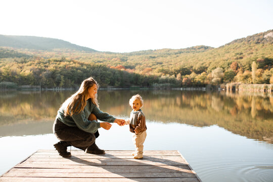 Young mother with little baby on wooden pier over autumn landscape view. Fall season. Motherhood.