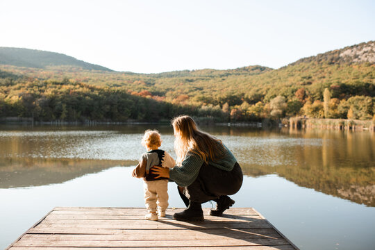 Young mother with little baby on wooden pier over autumn landscape view. Fall season. Motherhood.