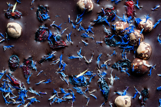Top view macro texture of dark chocolate with berries and cornflower. Full frame closeup background of premium dark chocolate bar topped with hazelnuts dried cranberries and blue cornflower petals