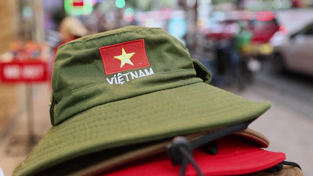 Green bucket hat with Vietnam flag and text embroidery displayed at souvenir shop in Nha Trang. Close up of military style sun hat on stall counter with blurred street background and traffic.
