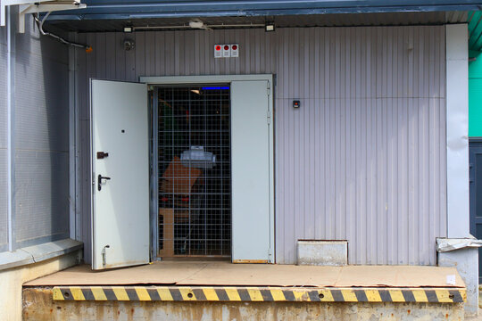 Open doorway reveals storage interior behind protective grid; weathered platform and hazard stripes emphasize industrial functionality and safety protocols.