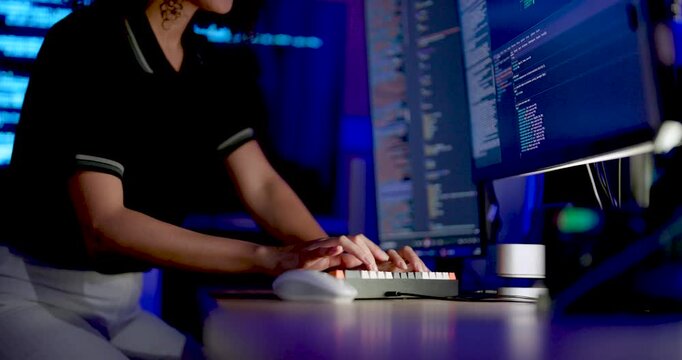 Adult female technology analyst types on keyboard, reaches toward monitor, and reviews source code across multiple screens during digital system monitoring and cybersecurity workflow at workstation.