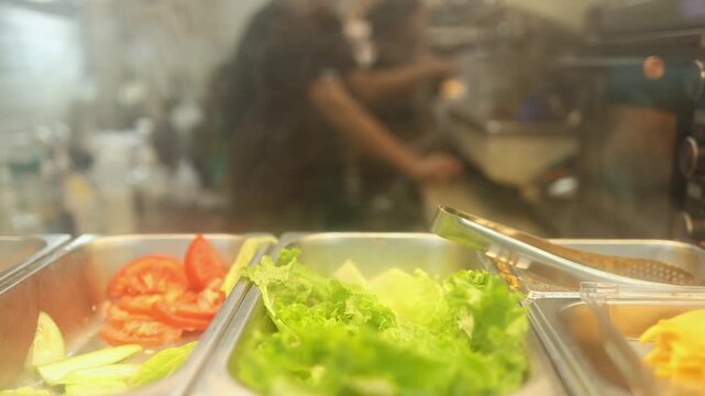 Fresh vegetables and salad ingredients in metal containers at local food eatery in Nha Trang. Close up of sliced tomatoes, cucumbers and lettuce on counter with blurred background cook.