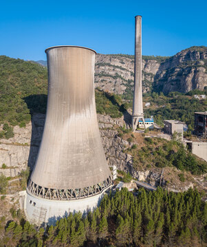 Derelict coal-fired power plant with large concrete cooling tower and tall chimney stack nestled in steep rocky mountain gorge, surrounded by green forest under clear blue sky