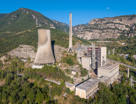 Derelict coal power plant with hyperbolic cooling tower, tall chimney, and partially demolished buildings nestled in pine forest between rocky mountain cliffs under clear blue sky