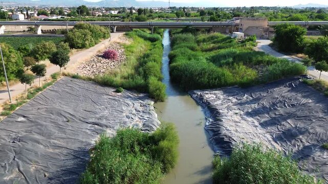 Aerial view of the Tajo Segura water transfer aqueduct crossing over a muddy river surrounded by green vegetation black plastic bank protection and agricultural fields under bright sunny sky