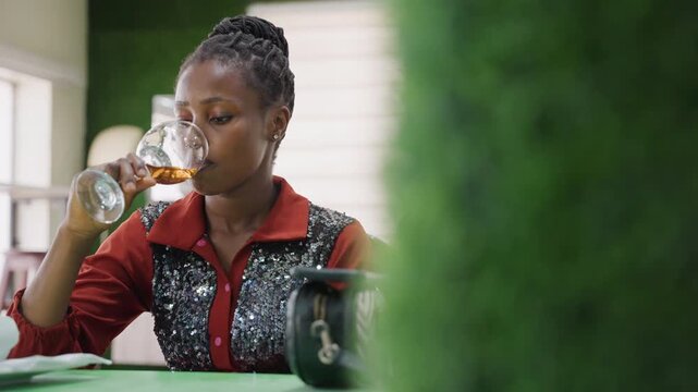 Black woman sipping wine alone, cozy cafe setting with green divider foreground, braided hairstyle, red blouse with sequins, amber wine in glass, thoughtful gaze, cinematic bokeh, natural window