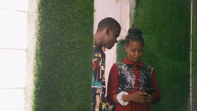 Black couple checking phone on date by green wall, young student and artist exchanging texts, girl in sequined dress, boy in colorful patterned shirt, shy smiles, close conversation, tender exchange,