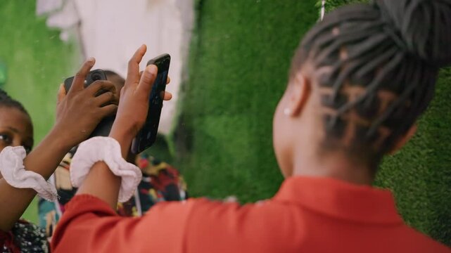 Woman taking photo with phone, closeup back view in red jacket capturing friend portrait against green backdrop, braided hair details, soft smiles and shy glances, intimate playful rehearsal with warm