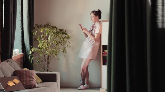 Young woman using smartphone by window in modern home interior. Concept of remote work communication, mobile productivity, flexible home office connectivity.