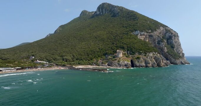 Aerial view of an ancient stone watchtower sits atop rugged rocky cliffs along a lush green coastline, overlooking vibrant turquoise Mediterranean sea. It is located on Mount Circeo, in Lazio, Italy.