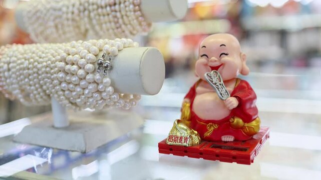 White pearl bracelets on display stand and laughing Buddha figurine at souvenir shop in Nha Trang. Close up of pearl jewelry and traditional solar-powered monk statue on glass counter.