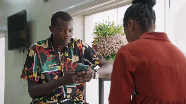 Black couple at cafe exchanging gift, man presenting box while woman smiles near window plant, warm sunlight, intimate conversation, playful laughter, close gestures, casual patterned shirt