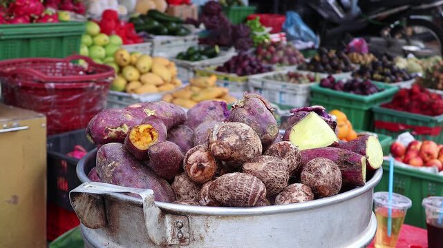 Fresh purple sweet potatoes cook in a steaming pot at an outdoor produce market. Steam rises from the hot container with colorful fruits and vegetables behind the food stall.