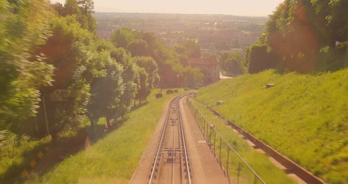 Funicular railway track run downhill between grassy slopes toward a distant Mondovi town in warm sunlight with a forward-moving ride perspective, Piedmont, Italy