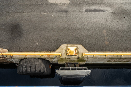 Top view of harbor dock edge with mooring bollard and tire fender above water.