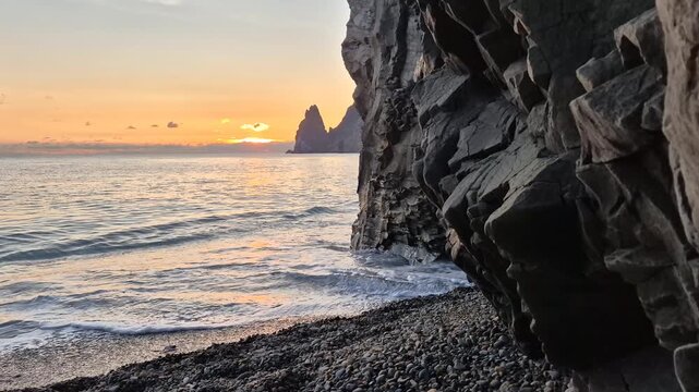 Sea sunset rocks, ocean waves crashing on pebble beach at golden hour next to dramatic rocky cliff landscape, tranquil nature
