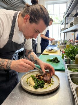 Professional tattooed male chef with a man bun carefully plating a gourmet plant-based vegan meat dish with broccolini and puree in a bright commercial restaurant kitchen.