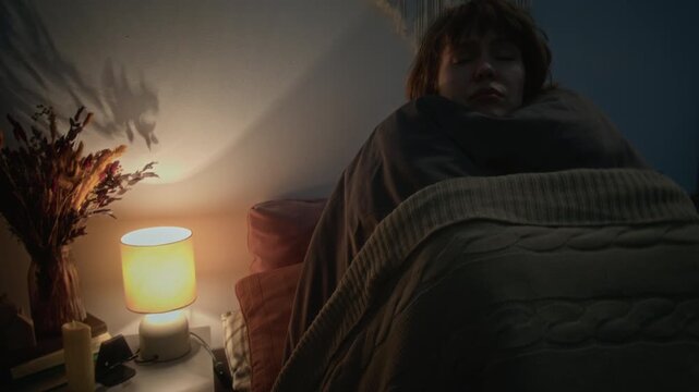 Young Caucasian woman turning on bedside lamp, wrapping in blanket, looking anxious as emergency lights flash through window. Ideal for mental health awareness or security system ads